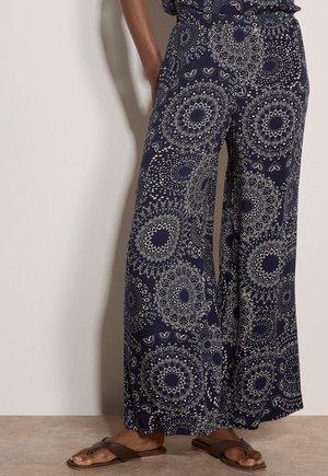 Marineblaue Hosen mit weitem Bein und weißen geometrischen Mandala-Mustern, getragen mit braunen Sandalen, vor einer hellen Wand stehend.