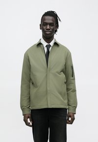 Olive green jacket with a zippered pocket on the sleeve, featuring a sleek, minimalist design and a smooth, textured fabric.