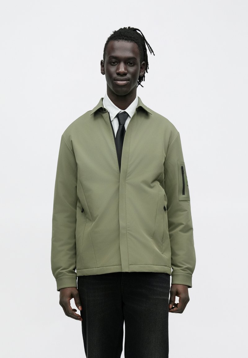Olive green jacket with a zippered pocket on the sleeve, featuring a sleek, minimalist design and a smooth, textured fabric.