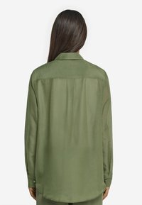 Chemise vert olive à manches longues avec une texture douce et lisse, poignets boutonnés, et un unique pli au dos, présentant un col classique.