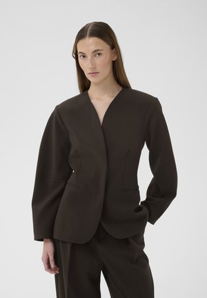 Pikkade juustega naine, kes kannab tumedat kohandatud bleiserit ja sobivaid pükse, seisab ühevärvilise heledat halli tausta ees.