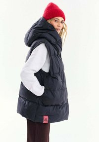 Žena so červenou pletenou čiapkou, čiernou vatovanou vestou s kapucňou, bielym tričkom s dlhým rukávom a tmavými nohavicami, pozerajúca sa cez rameno.