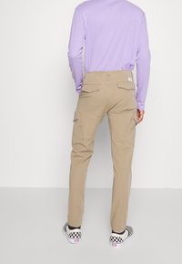 Beige cargobroek met meerdere zakken, katoenen materiaal, rechte pasvorm en zichtbare stikdetails. Gecombineerd met zwart-witte geblokte schoenen.