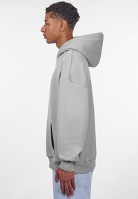 Grå hættetrøje lavet af blødt stof, med klassisk design, oversized pasform, frontlomme og ribbede manchetter. Hætten kan justeres.