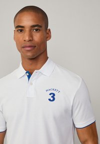 Λευκό polo μπλουζάκι με μπλε λεπτομέρειες και κεντημένα "HACKETT" και "3" στο στήθος. Διαθέτει δομητό γιακά και κοντό μανίκι.