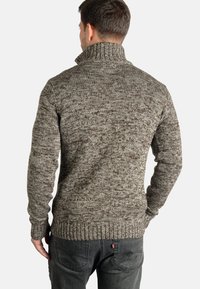 Knit sweater in a brown and cream heather pattern, featuring a high collar, ribbed cuffs, and a snug fit. Worn with dark gray jeans.