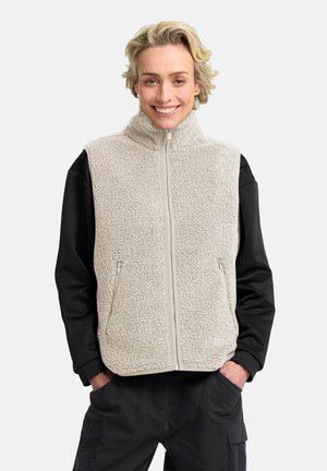 Gilet en polaire blanc cassé avec un col haut, une fermeture éclair intégrale et deux poches latérales ; associé à des manches longues noires et un pantalon foncé.