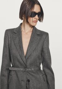 Frau mit kurzen braunen Haaren, die schwarze Sonnenbrillen und einen taillierten grauen Blazer mit breiten Revers trägt, vor einem schlichten hellen Hintergrund.