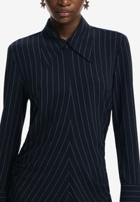 Rochie cambrată de culoare albastru marin, cu dungi verticale albe, guler ascuțit și mâneci lungi. Prezintă un închidere frontală cu nasturi.