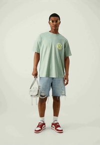 Light green cotton t-shirt with a graphic design, paired with distressed denim shorts and red and white sneakers. Clear bag and sunglasses.
