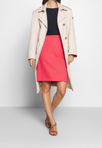 Trench coat beige chiaro con bottoni grandi, indossato sopra un top fitted blu navy e una gonna rosa. Gli accessori includono sandali rossi con cinturini.