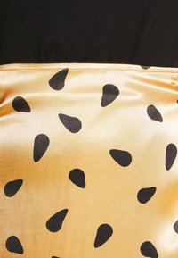 Top noir avec un panneau en satin doré brillant présentant des motifs de gouttes noires irréguliers, créant un contraste texturé entre les couleurs et les matériaux.