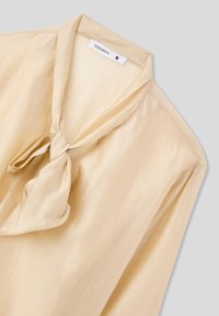 Blusa beige con dettaglio di fiocco al colletto, realizzata in tessuto leggero e morbido. Caratterizzata da maniche lunghe e un design dalla vestibilità comoda.