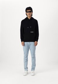 Sweatshirt noir à capuche avec une poche frontale et un logo, associé à un jean bleu clair et des baskets blanches ; accessoirisé avec des lunettes de soleil noires.
