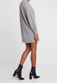 Robe en maille grise avec un col haut, des manches longues et des poches latérales. Associée à des bottines noires à bout pointu et à talon épais.