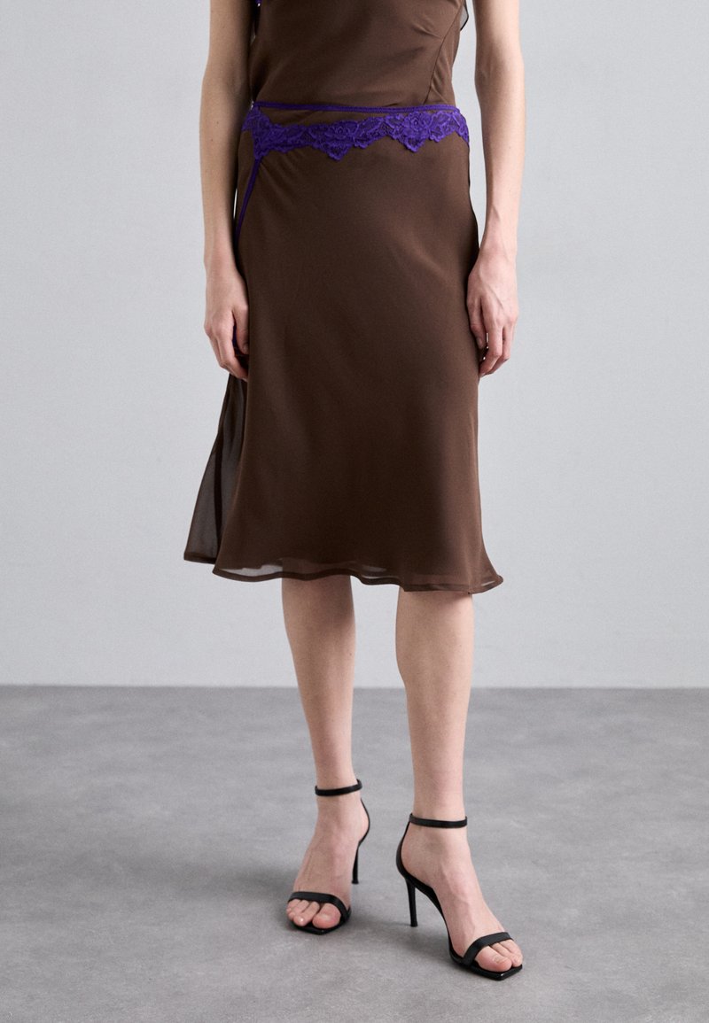 Abito lungo marrone con rifiniture in pizzo viola in vita. Tessuto liscio, orlo alla lunghezza del ginocchio, abbinato a tacchi neri con cinturini e un design minimalista.