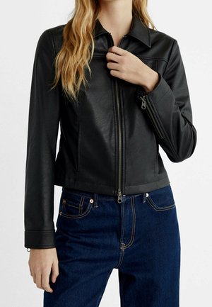 Femme portant une veste en cuir noire ajustée avec une fermeture éclair devant et un jean bleu en denim, main près de la fermeture éclair, longs cheveux blonds tombant sur l'épaule.