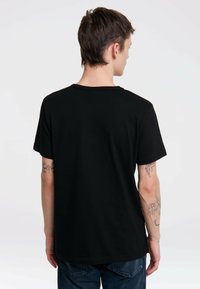 Camiseta de algodón negra con mangas cortas, cuello redondo y dobladillo recto. Incluye tatuajes visibles en los brazos, mostrados desde atrás.