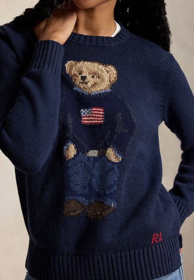 Tamsiai mėlynas megztinis su megztu meškiuku, dėvinčiu megztinį su Amerikos vėliava ir džinsais, RL logotipu prie apačios, dėvimas žmogaus su ranka ant kaklo.