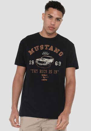 Young man wearing black T-shirt with a vintage Ford Mustang car graphic, text "MUSTANG 1969" and "THE BOSS IS IN" in brown and white lettering.
