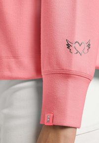 Różowy sweatshirt z miękką teksturą, prążkowanymi mankietami i srebrnym nadrukiem w kształcie serca z skrzydłami. Zawiera małą metkę z logo na rękawie.
