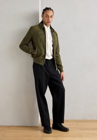 Giacca in suede verde oliva con colletto alto, chiusura con zip e due tasche laterali, abbinata a pantaloni neri larghi e stivali neri alla caviglia.
