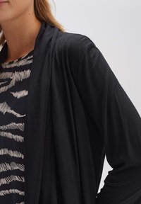 Zwarte open voorkaartigan van lichtgewicht stof, met een gedrapeerd ontwerp. Gedragen over een zwart-wit gestreept topje, dat de texturen benadrukt.