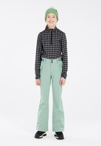 Haut zippé à motifs noir et blanc, pantalon vert menthe avec poches et boutons, porté avec des bottes noires, sur fond blanc.