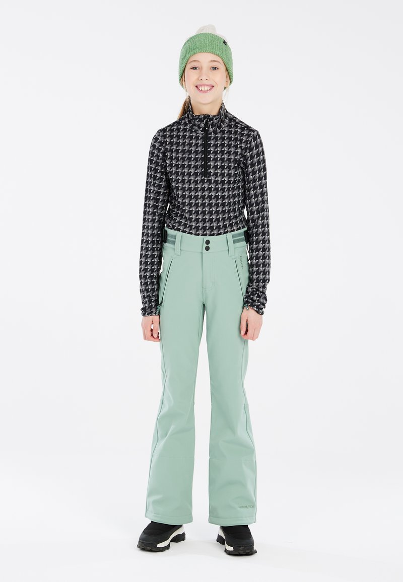 Haut zippé à motifs noir et blanc, pantalon vert menthe avec poches et boutons, porté avec des bottes noires, sur fond blanc.