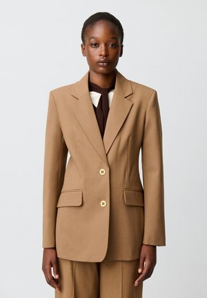 Donna che indossa una giacca da completo beige con due bottoni e pantaloni abbinati, in piedi davanti a uno sfondo grigio chiaro uniforme.