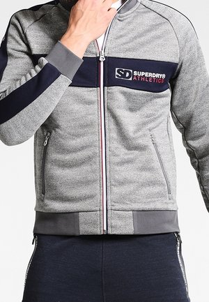 Mężczyzna noszący szarą kurtkę z zamkiem błyskawicznym z granatowym paskiem i logo „Superdry Athletics”, zapinający zamek jedną ręką.