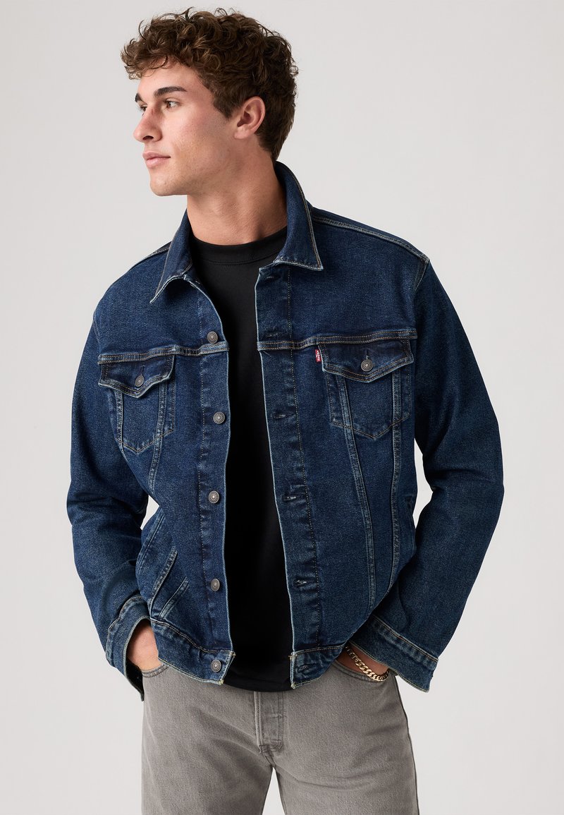 Veste en denim bleu foncé au style classique, à boutons sur le devant, avec deux poches poitrine et des accents en métal argenté. Associée à une chemise noire en dessous.
