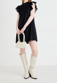 Mustat täplätunika, jossa on röyhelöhihat, yhdistettynä pieneen valkoiseen käsilaukkuun ja korkeisiin kermaisiin saappaisiin, neutraalin taustan edessä.