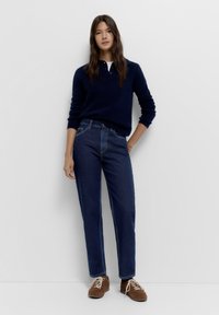 Nainen seisoo valkoista seinää vasten, yllään tummansininen pitkähihainen villapaita, tummansiniset farkut ja ruskeat tennarit, joissa on valkoiset nauhat.