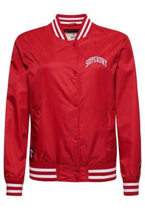 Person står op ad en hvid murstensvæg iført en rød Superdry-jakke, grå skjorte, sorte bukser og sorte og hvide sneakers.