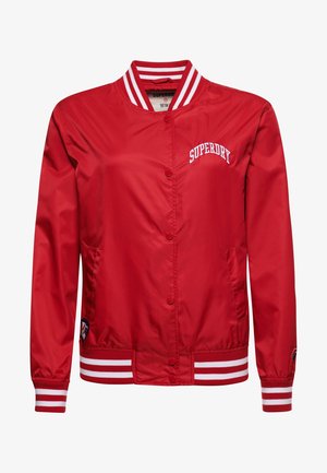 Person står op ad en hvid murstensvæg iført en rød Superdry-jakke, grå skjorte, sorte bukser og sorte og hvide sneakers.
