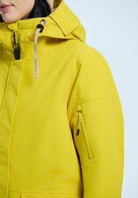 Giacca isolante gialla con colletto alto, tasca laterale con zip e accessori in contrasto. Il tessuto appare liscio con una finitura opaca.