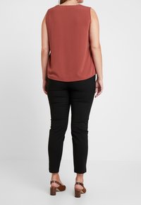 Top sans manches de couleur rouille avec un col arrondi, associé à un pantalon noir ajusté et des sandales à talons épais marron. Tissu lisse et coupe ajustée.