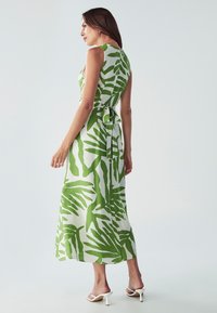 Maksi haljina od bijelog platna s uzorcima zelenog lišća; bez rukava, otvorenih leđa, s detaljem vezanja u struku, nosi se uz bijele sandale s visokom petom.
