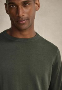 Grön stickad tröja med rundad halsringning och långa ärmar. Tyget verkar slätt med en medeltjock struktur. Ljus bakgrund.