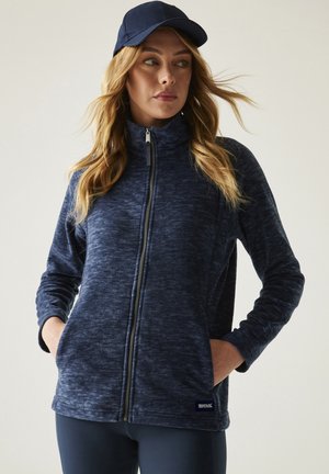 Kobieta w granatowej polarowej kurtce na suwak i pasującej czapce, ręce w kieszeniach, patrząca na bok na jednolitym tle.