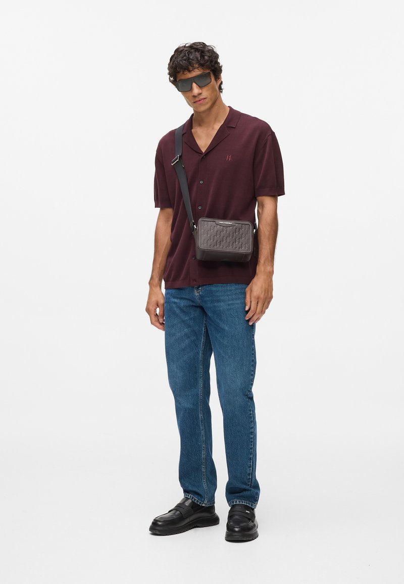 Sötét szürke, szőtt textúrájú, keresztpántos táska, téglalap alakú, állítható pánttal és kiemelkedő fém díszítéssel. A modellen kényelmes viselet látható.