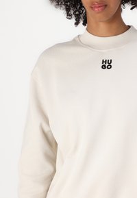 Majica boje kreme s okruglim izrezom, dugim rukavima i mekanom teksturom. Ima crni izvezeni logo "HUGO" na prsima.