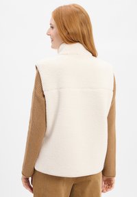 Crèmekleurige fleece vest met hoge kraag, voorzien van een gestructureerd oppervlak en een rugpand. Gepaard met een bruine gebreide trui.