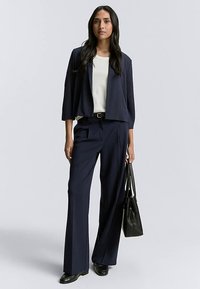 Marineblazer und passende, weit geschnittene Hose, kombiniert mit einem weißen Oberteil. Accessoires sind eine schwarze Handtasche und schwarze Schuhe. Glatte Stoffstruktur.