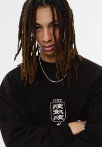 Fiatal személy hosszú rasztahajasztással, fekete pulóvert viselve, amelyen három oroszlános címer található, ezüstlánccal a nyakában, karjait keresztbe fonta, semleges arckifejezéssel.