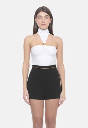 Donna con capelli castani lisci che indossa un top bianco senza spalline con dettaglio al collo e pantaloncini neri a vita alta con sottile cintura a catena dorata.