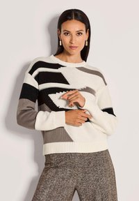 Vrouw in een gebreide trui met zwart, wit en grijs geometrisch patroon en textuurbruin herringbone broek.