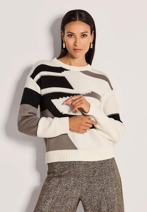 Frau trägt einen Strickpullover mit schwarz-weißen und grauen geometrischen Mustern sowie einer strukturierten braunen Fischgrät-Hose.