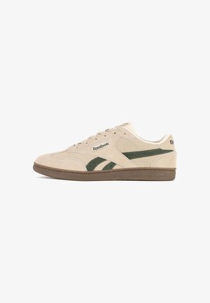 Beige suede sneakers with green accents, perforated design, flat brown rubber sole, and lace-up closure. Features Reebok logo on the side.